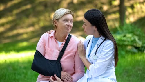 An older man in a shoulder sling recovering at home with a caregiver nearby